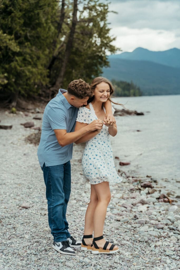 Joyful couple laughing together along the rocky shoreline of Lake McDonald during a romantic engagement session in Glacier National Park with mountain lake views and documentary style photography